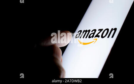 Amazon logo on a smartphone screen in a dark room and a finger pointing at it. Stock Photo