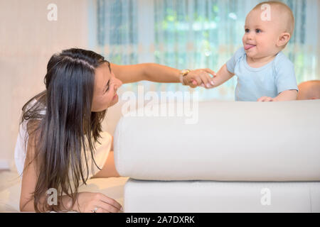 Cheerful mom is enjoying playing with her son while they are located on couch. She is pointing fingers at kid with fun and little boy is watching her Stock Photo