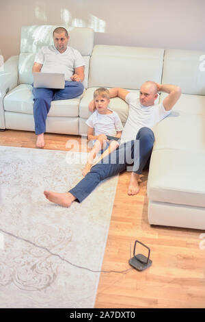 young family watching television, two guys and a little boy sitting on the sofa and the floor in the living room at home. Stock Photo