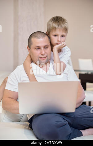 The son and his father are sitting on the couch, looking at a laptop, a happy family. Stock Photo