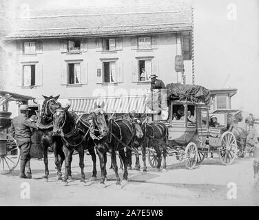 An early vintage black and white photograph showing a stage coach pulled by six horses being prepared to leave a small European town with passengers on board. The stage coach driver sits on top with his legs covered by a blanket. Some passengers are in the enclosed part of the coach, others sit in the outside exposed part of the coach.