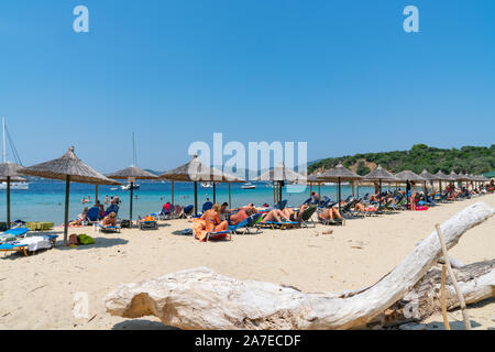 Skiathos Greece - August 1 2019; Beachgoers relaxing under sun umbrellas on idyllic Greek Island beach with large piece driftwood in foreground Stock Photo