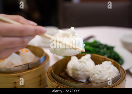 A womans hands holding chopsticks picking up a bbq pork bun out of a bamboo steamer at a dim sum restaurant. Stock Photo