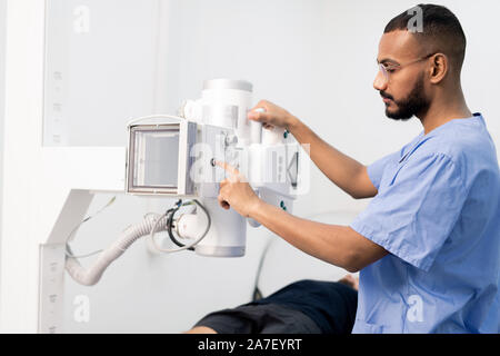 Young mixed-race man in blue uniform pressing button on new medical equipment Stock Photo