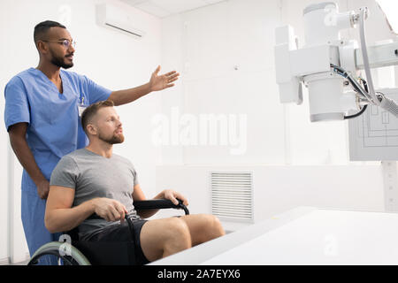 Sportsman sitting on wheelchair by medical equipment while doctor pointing at it Stock Photo