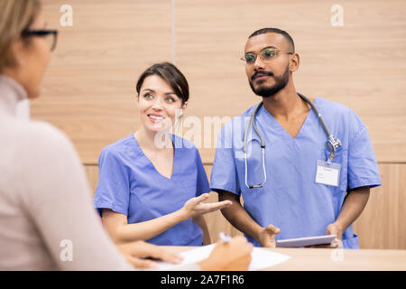 Pretty clinician in uniform smiling at patient while introducing her colleague Stock Photo