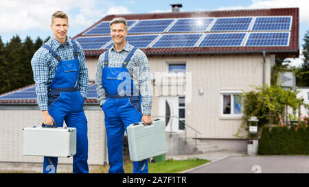 Portrait Of Smiling Electricians Holding Tool Boxes Standing In Front Of House With Solar Panel Stock Photo