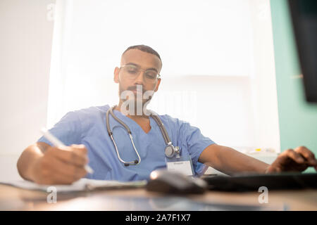 Bearded serious doctor or intern in uniform making working notes by table Stock Photo
