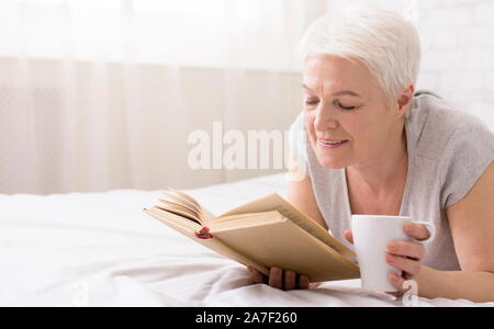 Calm senior woman lying on bed with book and coffee cup Stock Photo