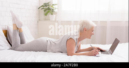 Cheerful senior lady chatting on laptop online, lying on bed Stock Photo
