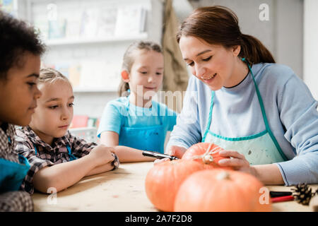 Happy young woman surrounded by schoolkids drawing face on pumpkin Stock Photo