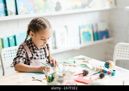 Cute casual schoolgirl with paintbrush drawing Christmas picture by desk Stock Photo