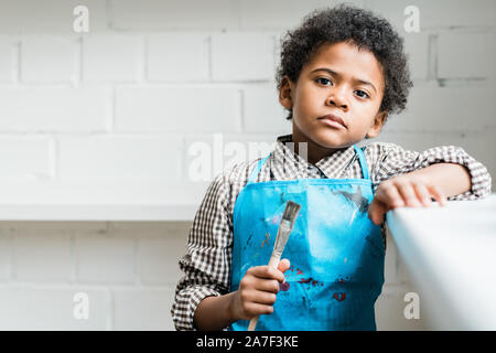 Serious African schoolboy in blue apron holding paintbrush in hand Stock Photo