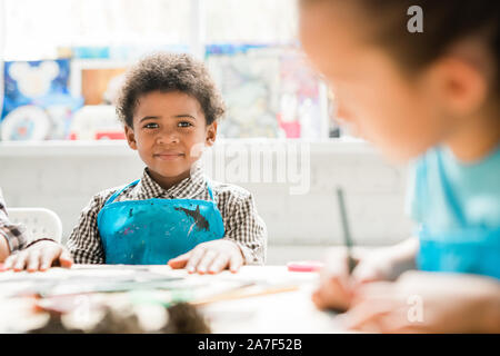 Cute schoolboy of African ethnicity in blue apron sitting by desks at lesson Stock Photo