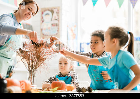 Teacher and schoolkids in aprons putting handmade paper bats on dry branches Stock Photo