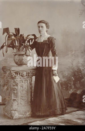 Late Victorian or early Edwardian cabinet photo card portrait of young Edwardian woman holding a letter and wearing a sombre, dark dress, posing next to potted plant - an aspidistra which were popular plants at this time. circa 1901, Truro, Cornwall, U.K. Stock Photo