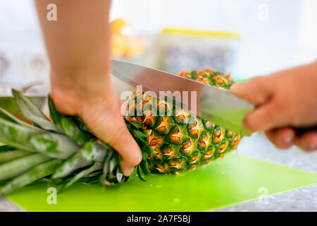 Woman hands with knife cutting pineapple top on cutting board in domestic kitchen. Close-up. Stock Photo
