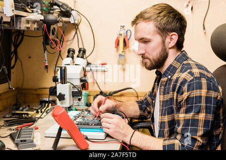 Young bearded mechanic with two soldering-irons sitting by workplace in workshop Stock Photo