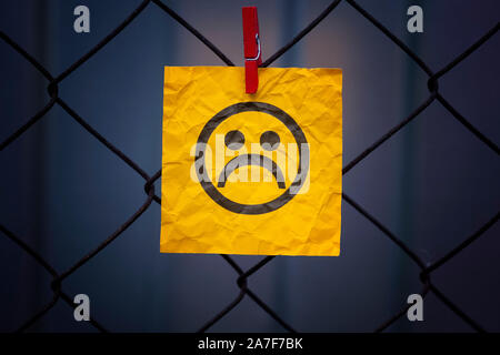 Yellow paper note with sad face hanging on a metal mesh fence. Close up. Stock Photo