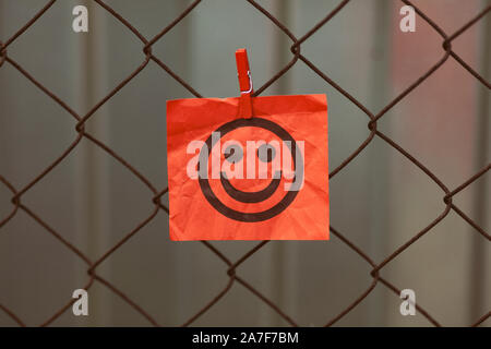 Paper note with happy face hanging on a metal mesh fence. Close up. Stock Photo