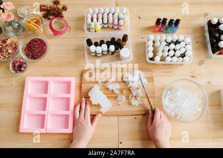 Young woman holding knife over crystals of soap mass while cutting it on board Stock Photo