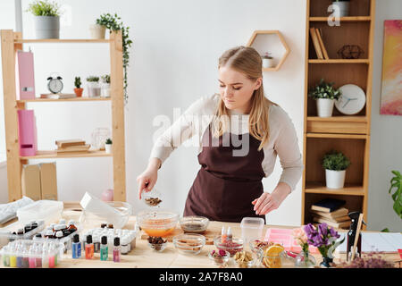 Young craftswoman adding grated dry spices into bowl with liquid soap mass Stock Photo