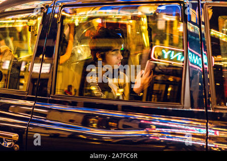 A young male checks his smart phone whilst sitting in the back of a London Taxi on neon illuminated evening Stock Photo