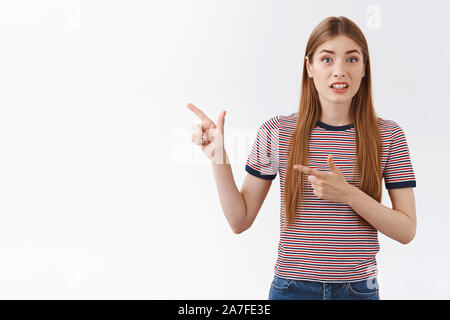 Alarmed and worried young girl with long hair feeling anxious about doubtful strange action happening, pointing fingers left, squinting and cringe Stock Photo