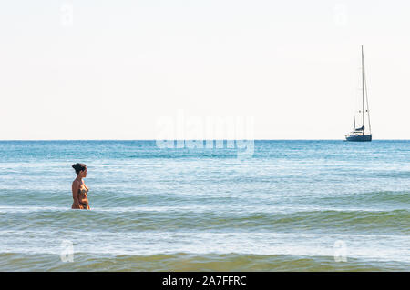 Tenda Gialla, Province of Grosseto, Tuscany, Italy - September 03, 2019: Young woman standing in the sea alone and looking on the sailing yacht far aw Stock Photo
