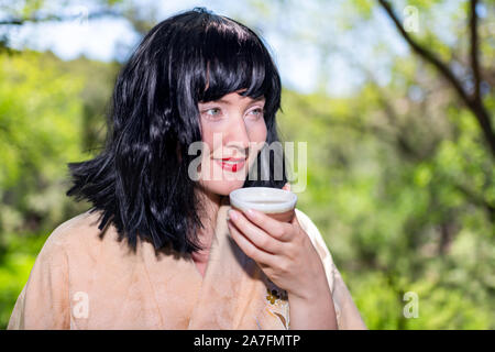 Young girl woman happy face closeup smiling holding drinking green tea cup outside in backyard garden with black hair Stock Photo