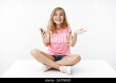 Cute silly pretty blond young daughter fool around joyfully playing mobile phone, sit crossed legs floor enjoy browsing internet, shrug raise hand Stock Photo