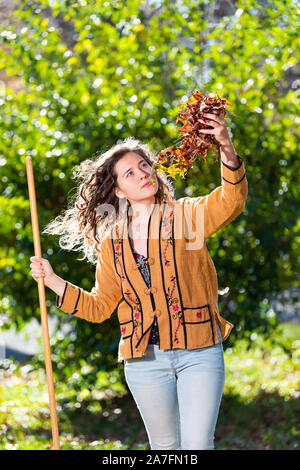 Young woman homeowner in yard backyard raking collecting of dry autumn foliage oak leaves standing holding foliage with rake in sunny fall Stock Photo