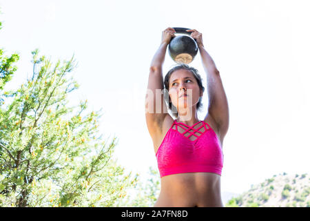 Young fit woman with abs muscles on stomach lifting heavy kettlebell doing exercise in outdoors outside park holding weight Stock Photo