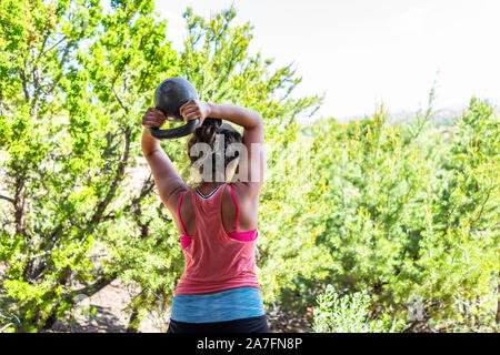 Back of young fit woman with heavy kettlebell doing triceps exercise and muscles in outdoors outside park holding weight lifting Stock Photo