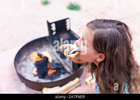 Young woman eating gooey roasted marshmallows smores with chocolate and rice cake cracker by fire in campground campfire grill Stock Photo