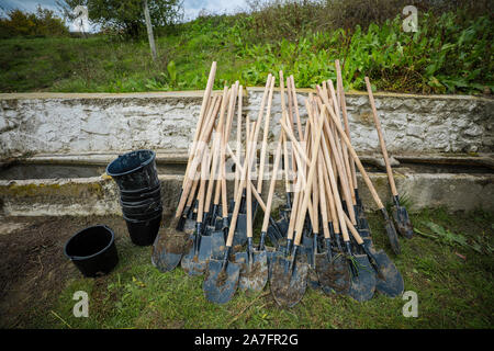 Dirty plastic buckets and metal shovels with wood handles on the grass, near a rock watering spring in a rural area after people planted trees. Stock Photo