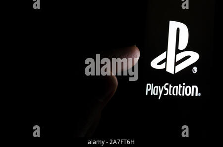PlayStation logo on a smartphone screen in a dark room and a finger touching it. Stock Photo