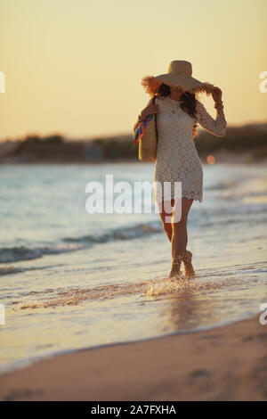 Full length portrait of trendy middle age woman in white dress and straw hat on the seacoast at sunset having fun time. Stock Photo