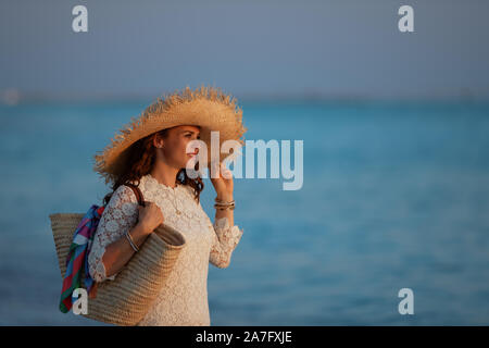 Side view of smiling young woman in white dress and straw hat on the ocean shore at sunset looking into the distance.