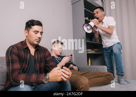 Young man shouting through a megaphone at friends which using a mobile phone. Stock Photo