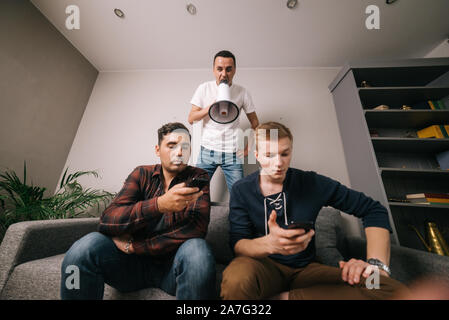 Young man yelling through a loudspeaker at friends watching TV. Stock Photo
