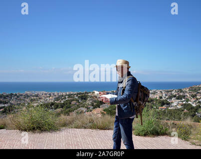 Traveler man on a viewing platform on top of a mountain with a map of the area in his hands, against the backdrop of a city by the sea Stock Photo