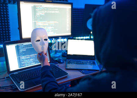 Hacker in hood holds mask in hand, crime lifestyle Stock Photo