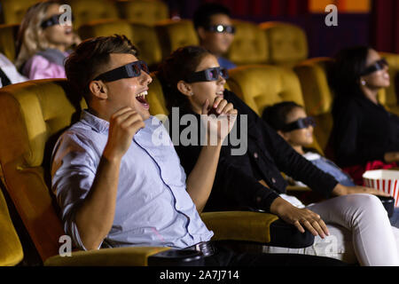 Amazed young man and woman couple in 3D glasses sitting on comfortable chairs and watching exciting film in movie theater being on date Stock Photo