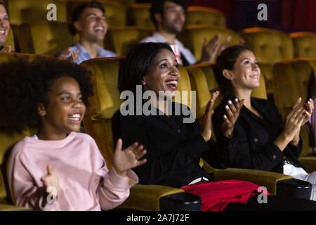 Excited black woman and girl audience smiling and clapping hands while sitting amidst people after movie in cinema Stock Photo