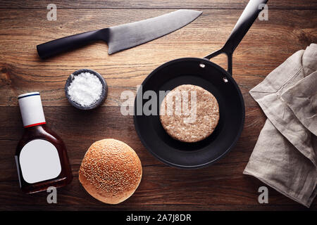 Raw burger meat patty in a frying pan, decorated with salt, sauce, bread, knife and napkin on a wooden table background. Hamburger preparation. Stock Photo