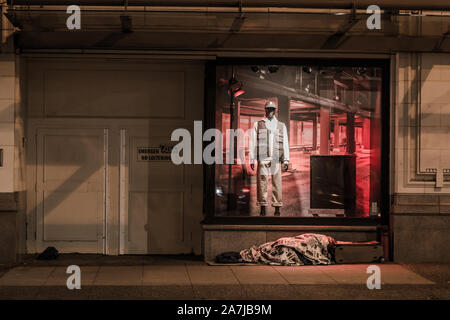 A jarringly ironic scene of a homeless man sleeping on the street right in front of a clothing display of an affluent department store. Stock Photo