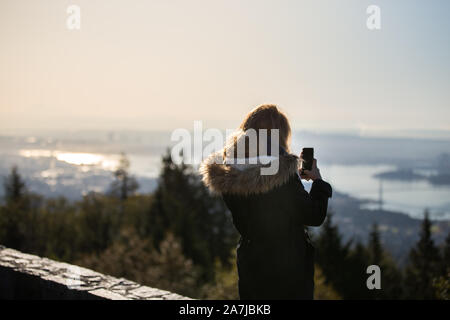 A young woman in a winter coat at the Cypress Mountain lookout taking a photo of downtown Vancouver on her cell phone. Stock Photo