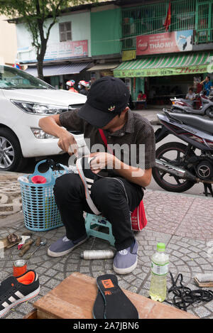 A young Vietnamese man, wearing a black baseball cap, sitting on wooden stool on sidewalk repairing a pair of black sneakers. Stock Photo
