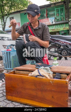 A young Vietnamese man, wearing a black baseball cap, sitting on wooden stool on sidewalk repairing a pair of black sneakers. Stock Photo